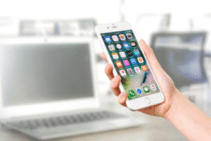 A hand holds a smartphone displaying app icons in front of an open laptop on a blurred office desk.